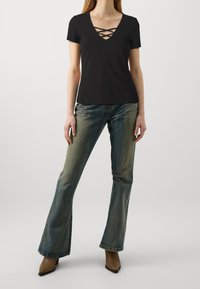 Schwarzes, tailliertes T-Shirt mit V-Ausschnitt und Kreuzdetail am Ausschnitt, kombiniert mit ausgestellten blauen Jeans und beigen spitzen Stiefeln.