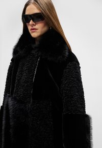 Melnas pārmērīgi lielas mētelis ar teksturētu ādas dizainu, ar pūkainu apkakli, ar rāvējslēdzēja aizdari un spīdīgiem mīkstiem auduma akcentiem. Valkā melnas saulesbrilles.