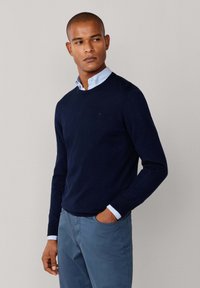 Marineblauwe crewneck trui van een zachte brei, met een subtiele logo en gelaagd over een lichtblauw overhemden. Ggedragen met lichtblauwe broek.