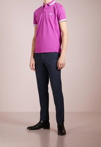 Chemise polo violette avec col rayé, associée à un pantalon sur mesure bleu foncé et des bottes noires, montrant une silhouette ajustée.