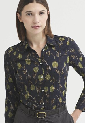 Jeune femme aux cheveux bruns lisses porte un chemisier bleu marine boutonné avec un imprimé floral vert et un pantalon gris avec une ceinture noire.