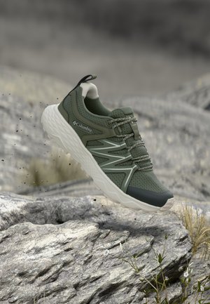 Olive green Columbia hiking shoe with white sole in mid-air above rocky ground in outdoor natural setting.