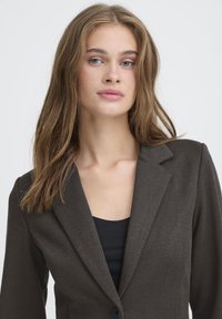 Blazer marrone a spina di pesce con chiusura a un bottone, rever a lancia e taglio sartoriale. Indossato sopra una maglia nera, con tessuto strutturato.