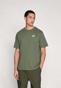 Λαδί βαμβακερό T-shirt με στρογγυλή λαιμόκοψη και κοντά μανίκια, με λευκό λογότυπο στο αριστερό στήθος. Απλός, χωρίς διακοσμήσεις σχεδιασμός.