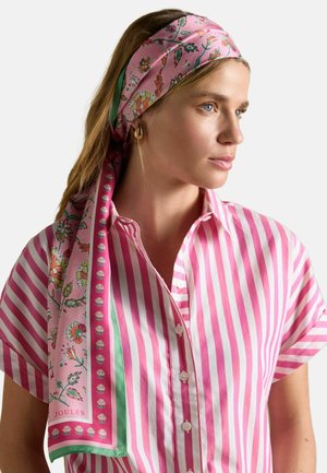 Frau mit blauen Augen, die ein rosa-weiß gestreiftes Hemd und ein pinkfarbenes Blumentuch trägt, schaut nach rechts vor einem schlichten Hintergrund.