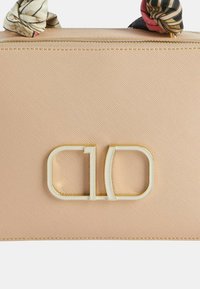 Borsa a tracolla beige con texture, dotata di hardware dorato e un logo bianco in evidenza. Presenta una sciarpa decorativa annodata al manico per un tocco di stile.