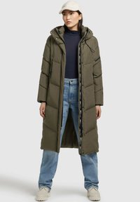 Lunga giacca a vento verde oliva con cappuccio, dotata di chiusura con zip, tasche laterali e cordini. Indossata sopra una maglia blu scuro e jeans blu.
