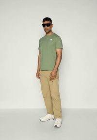Camiseta de algodón verde con logo, pantalones ligeros color beige con bolsillos, zapatillas blancas y gafas de sol negras. De pie contra un fondo simple.