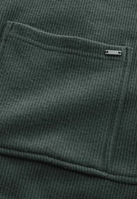 Tissu gaufré vert foncé avec une poche rectangulaire comportant une petite étiquette métallique "NEXT" dans le coin supérieur droit.