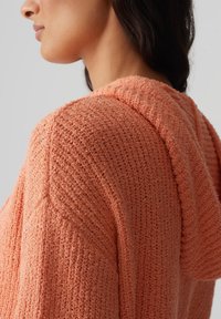 Maglione con cappuccio in tessuto lavorato di un arancione chiaro, caratterizzato da cuciture aperte e una vestibilità rilassata. Spalle accentuate e dettagli sul cappuccio.