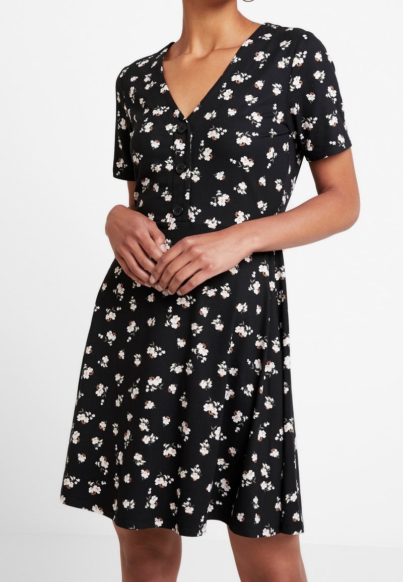 Femme portant une robe noire à manches courtes avec imprimé floral blanc et boutons devant, les mains délicatement jointes à la taille.