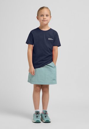 Marineblauses T-Shirt, hellgrüner Rock und türkisfarbene Sportschuhe mit schwarzen Akzenten. Das T-Shirt hat ein Logo auf der Brust.