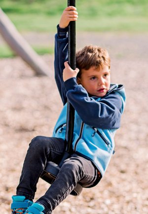 Child gripping a black pole, wearing a blue zip-up jacket with dark sleeves, black jeans, and turquoise shoes while sitting on a swing.