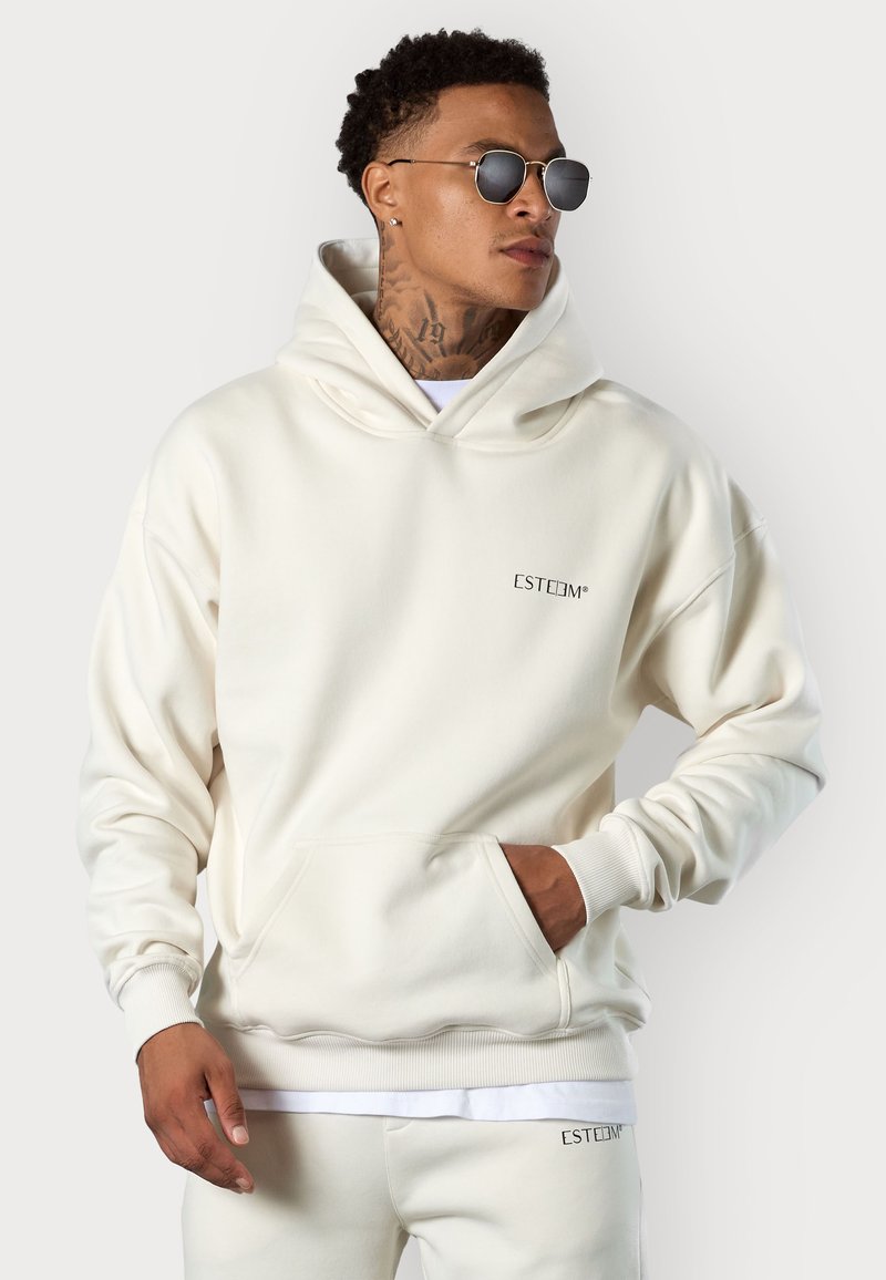 Sweat à capuche crème avec une grande poche avant, des poignets côtelés, une marque discrète sur la poitrine et une texture douce. Porté avec des lunettes de soleil.