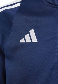 Tecido desportivo azul marinho com uma superfície texturizada, apresentando um logótipo de três riscas brancas e três picos paralelos bordados no peito.