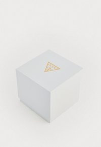 Caixa quadrada branca texturizada com um logótipo dourado triangular em cima, apresentando a marca "GUESS". Design simples, sem padrões ou acentos adicionais.