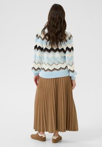 Femme aux longs cheveux ondulés portant un pull zèbre bleu, crème et marron, une jupe plissée marron et des baskets marron, vue de dos.