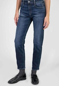 Vrouw in donkerblauwe jeans, grijze sokken en glanzende zwarte instappers, staand tegen een lichte achtergrond.