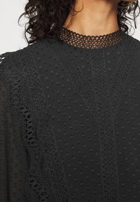 Musta tekstitoppi, jossa on kohokuvioituja pisteitä ja monimutkainen pitsireunus kaula-aukossa ja hartioilla. Pitsissä on kukkakuviota.