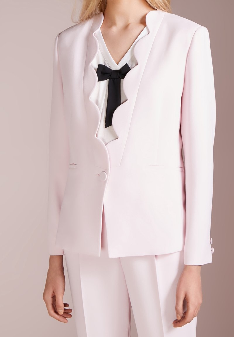 Vaaleanpunainen räätälöity bleiseri, jossa on aaltoilevat läpät, yksittäinen nappikiinnitys ja kaksi etutaskua. Materiaali näyttää sileältä ja jämäkältä.