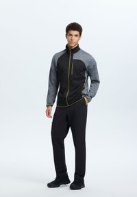 Giacca atletica nera e grigia con colletto alto, tasche con zip e dettagli verde oliva, abbinata a pantaloni e scarpe atletiche nere.