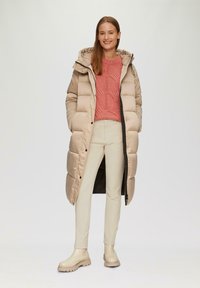 Manteau d'hiver beige avec capuche, pull en laine rose texturé, pantalon crème et bottes bégaie à semelle épaisse beiges. Tissu lisse, design longueur genou.