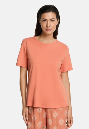 Mujer de cabello oscuro que lleva puesta una camiseta coral lisa de manga corta y pantalones coral con estampado, de pie frente a un fondo claro.