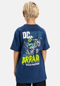 Námořnická modrá bavlněná tričko s živou grafikou jezdců motocrossu, textem "DC SHOE CO" a "PURE BRRAAAP" v jasně zelené a bílé barvě.