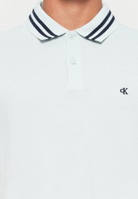 Hvit polo-skjorte med marineblå doble striper på kragen, to knapper, og liten svart "CK" logo på venstre bryst.