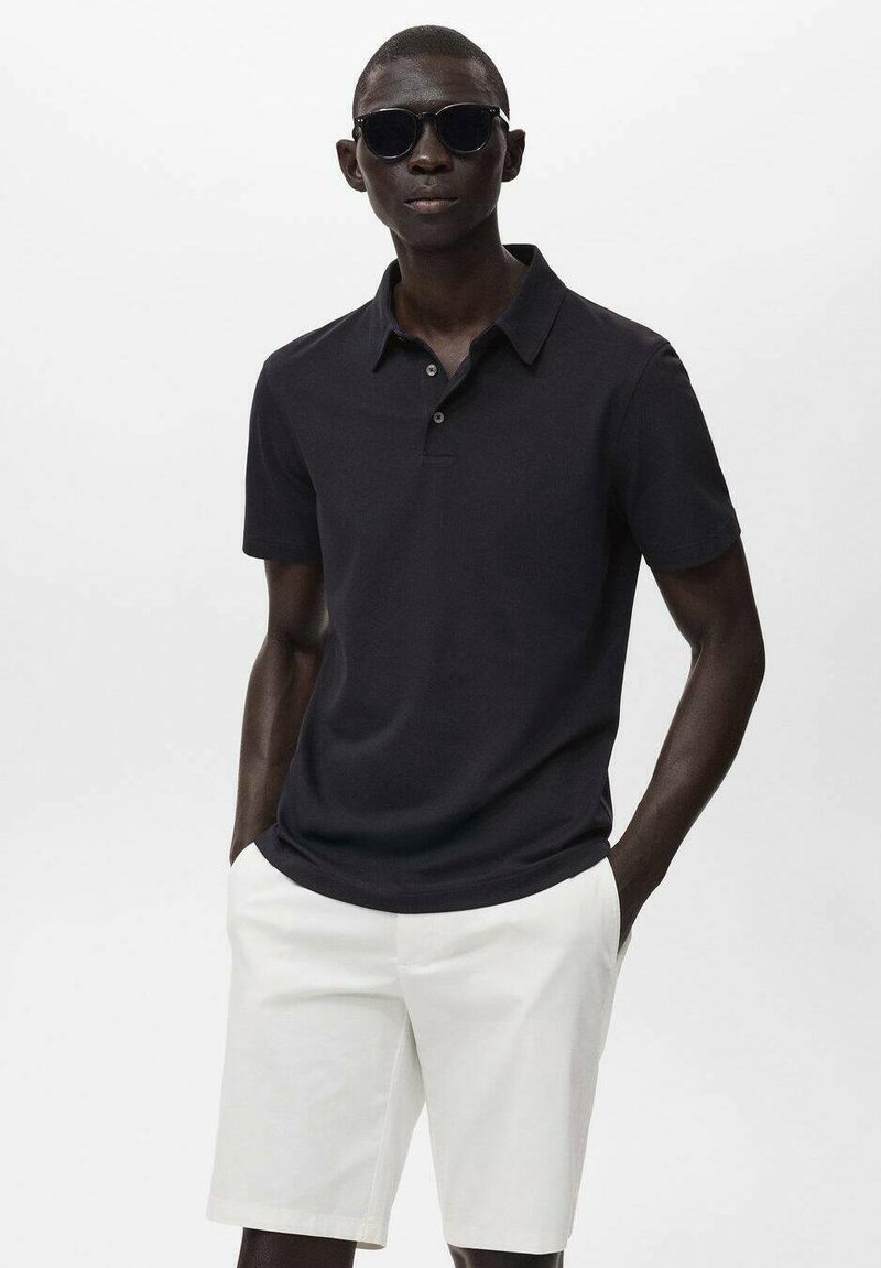 Svart polo t-shirt med krage och tre knappar, i kombination med vita shorts. Modellen bär mörka solglasögon och står mot en enfärgad bakgrund.