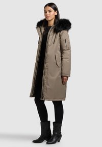 Pikk beež toonis turske talvejope musta kunstkarusnahast kapuutsiga, küljetaskud lukuga ja elastsete paeladega. Kandmisel musta riietuse ja saapade peal.