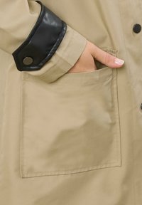 Giacca beige con un polsino in pelle nera con bottone a pressione e una tasca frontale. Il tessuto appare liscio con una leggera lucentezza.