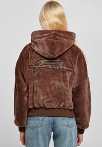 Haut à capuche en fausse fourrure marron avec un capuchon à cordon de serrage, des poignets en côte et un ourlet côtelé. Présente un logo de marque texturé en marron clair dans le dos.