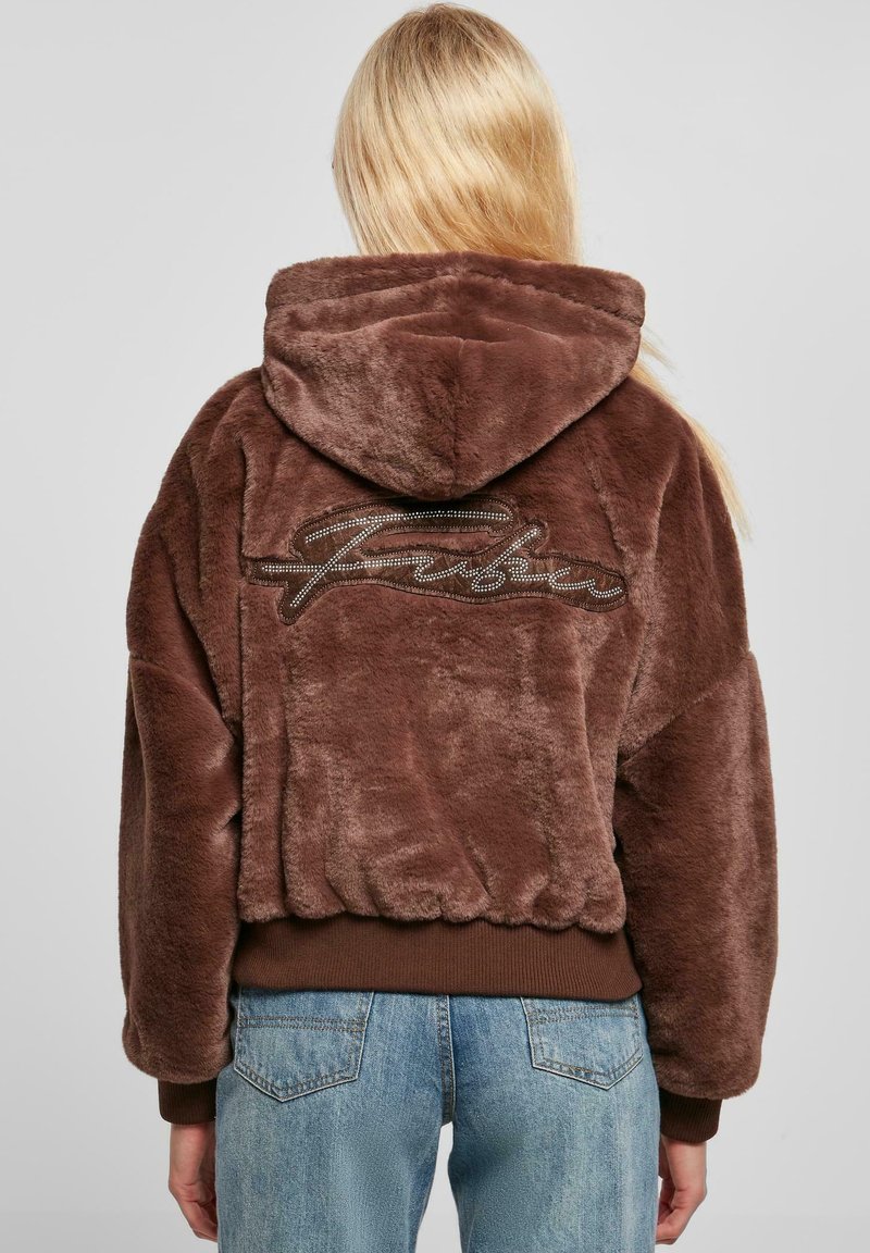 Haut à capuche en fausse fourrure marron avec un capuchon à cordon de serrage, des poignets en côte et un ourlet côtelé. Présente un logo de marque texturé en marron clair dans le dos.