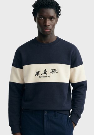 Giovane uomo che indossa una felpa blu navy e bianca con la scritta Barcelona '92 e icone di atletica leggera per corsa e ostacoli sul petto.