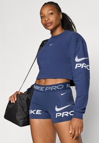 Top corto sportivo navy con logo sulla manica, abbinato a pantaloni corti navy con testo bianco "NIKE" e "PRO". Borsa da palestra nera con tracolla.