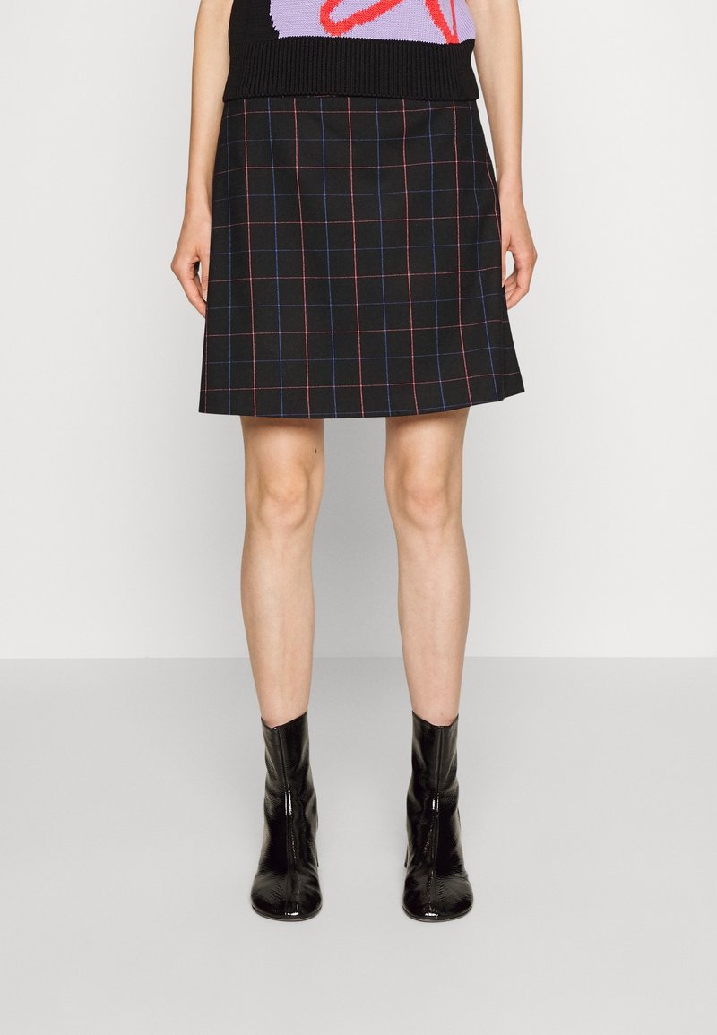 Jupe noire trapèze avec motif à carreaux rouges et bleus, portée avec des bottines noires vernies et un haut sans manches coloré.