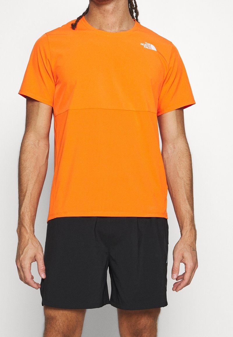 Camiseta atlética naranja con un patrón texturizado, mangas cortas, escote redondeado y un logo blanco en la parte superior izquierda del pecho. Combinada con pantalones cortos negros.