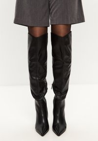 Botas de cuero negro hasta la rodilla con puntas en forma de flecha, acentos de cremallera en los laterales y un diseño ligeramente holgado en el eje superior.