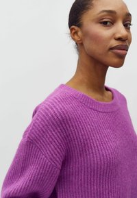 Junge Frau mit dunkler Haut, die einen strukturierten lila Pullover trägt und leicht seitlich vor einem schlichten weißen Hintergrund steht.