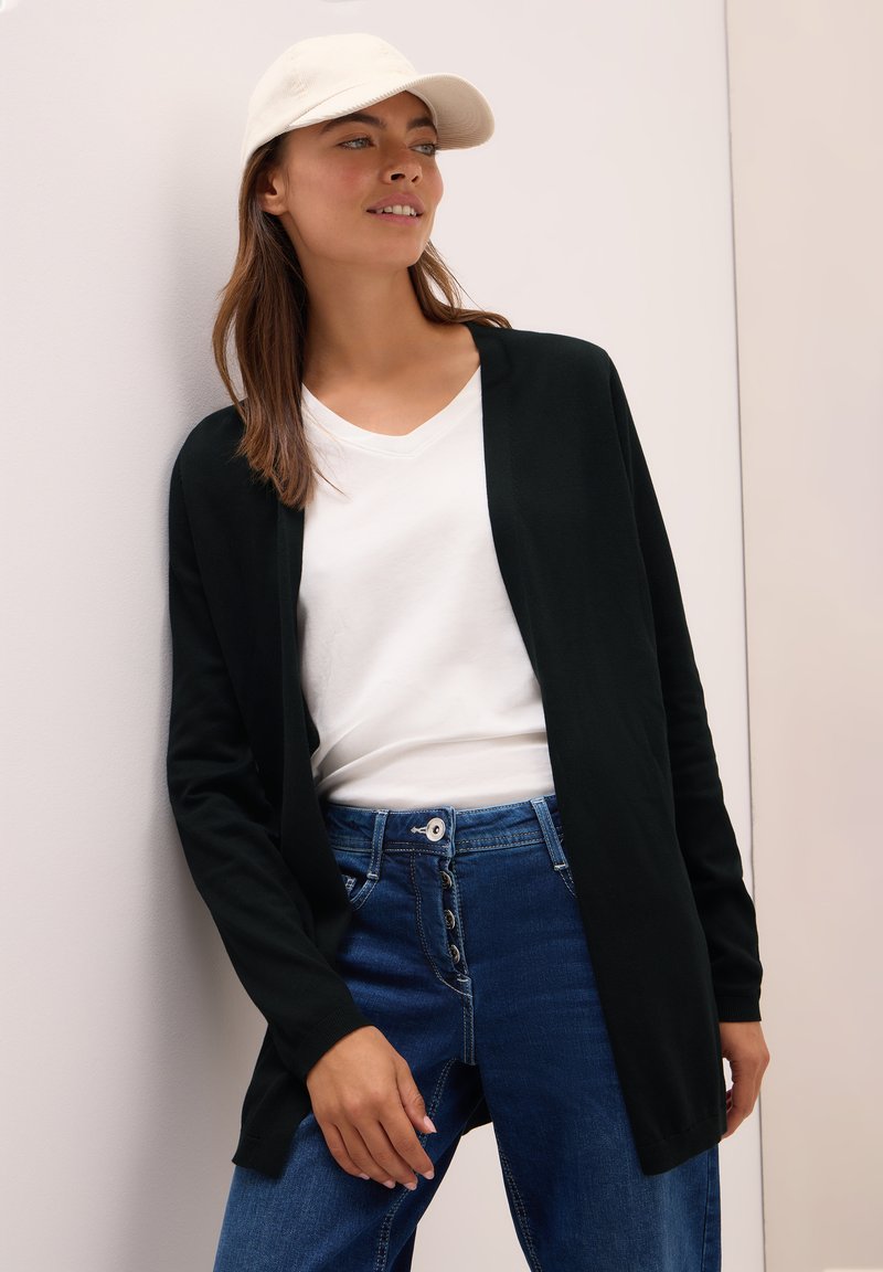Zwarte open voorkardigan over een wit T-shirt, gecombineerd met blauwe jeans met een knoopsluiting. Model draagt een crèmekleurige baseballcap.