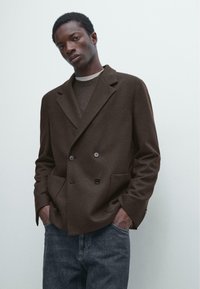 Brązowa wełniana marynarka dwurzędowa z dużymi guzikami, klapami z wycięciem i kieszeniami, zestawiona z jasnym swetrem i ciemnymi dżinsami.