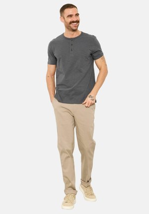 Lächelnder Mann mit Schnurrbart, der ein graues Henley-Shirt, beige Hosen und beige Sneaker trägt und mit einer Hand in der Tasche steht.