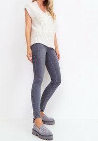 Vrouw in een mouwloze witte gebreide vest, grijze skinny jeans en grijze platform instappers, staand tegen een witte muur.