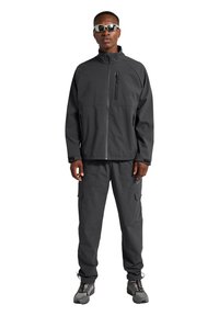 Schwarze Softshelljacke mit hohem Kragen und durchgehendem Brustreißverschluss, kombiniert mit passenden Cargo-Hosen, die seitliche Taschen und dezente Branding-Elemente aufweisen.