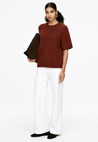 Rostfarbener kurzärmeliger Strickpullover, lockere Passform, kombiniert mit weißen weit geschnittenen Hosen und einer dunkelbraunen Clutch. Glatte Textur, keine Muster.