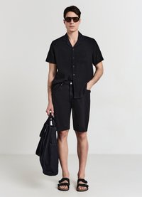 Camicetta nera a maniche corte con tessuto texture, pantaloni neri, occhiali da sole e sandali neri. Tiene una borsa a tracolla nera con manici.