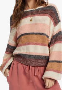 Strickpullover mit gestreiftem Muster in Rosa-, Braun- und Cremetönen, mit weitem Ausschnitt und geripptem Saum. Kombiniert mit pinken Jogginghosen.