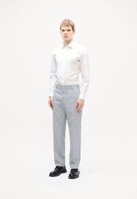 Chemise blanche à boutons et pantalon gris clair, avec une coupe droite. Le modèle porte des chaussures noires, se tenant devant un fond uni.