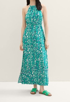 Robe maxi à col halter en turquoise avec des taches abstraites blanches et violettes. Tissu léger, taille élastique, ourlet superposé. Sandales plates vertes.